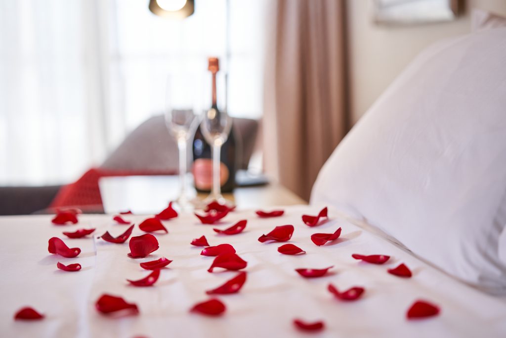 From above closeup of bed covered with white bedclothes and cushions with scattered petals of fresh red roses and bottle of champagne with glasses on bedside table in hotel room